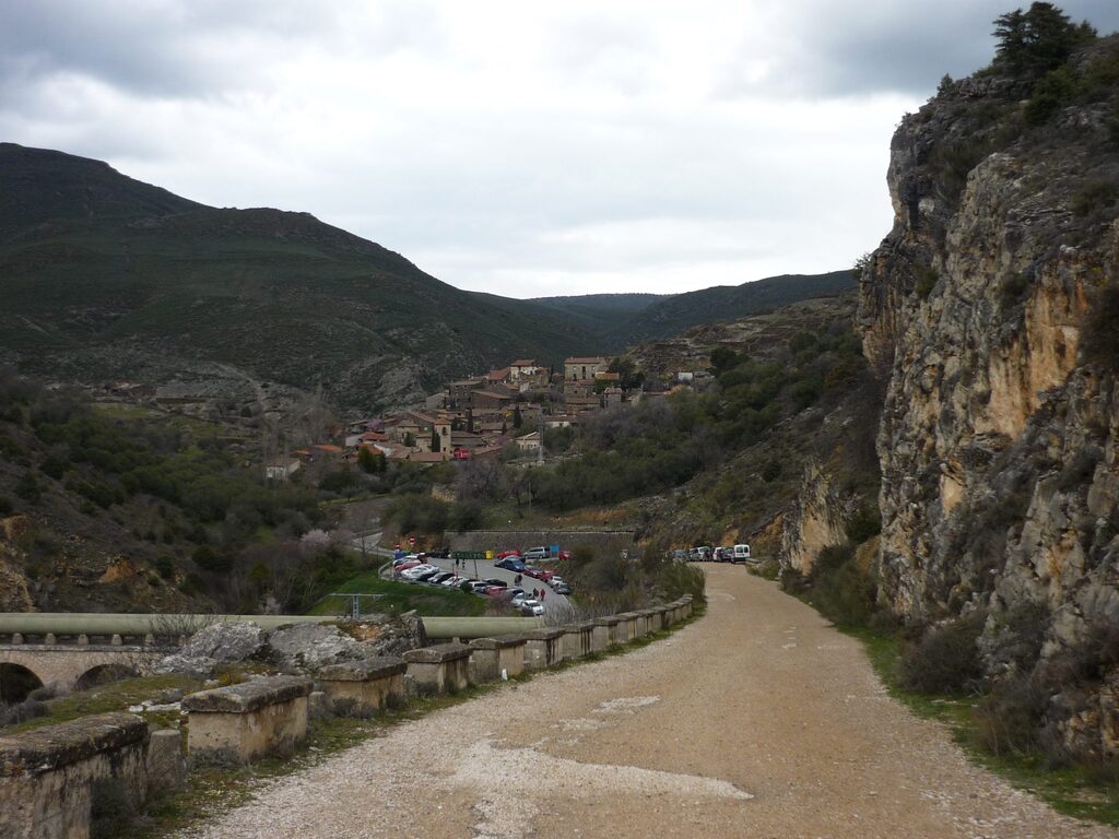 Camino de tierra en descenso en el monte, con una vista panorámica de un pueblo a lo lejos.